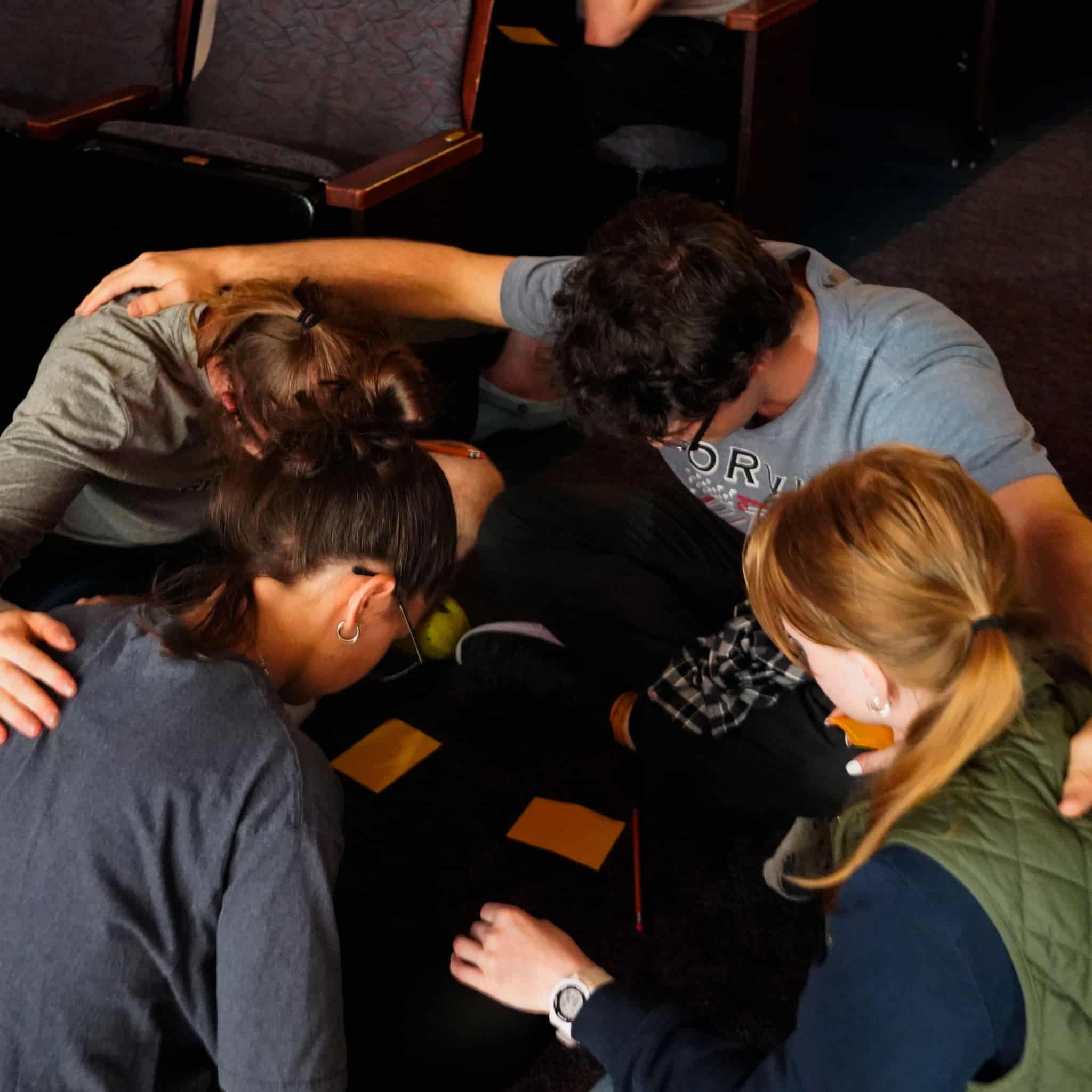 Four students sit on the floor in a prayer circle with their heads down