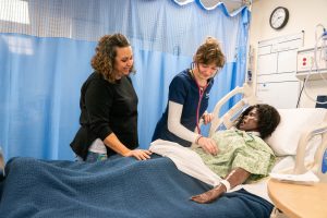 Nursing student using a stethoscope on a patient mannequin with instructor guidance in a clinical simulation lab