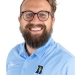 Professional headshot of a smiling bearded man with round glasses wearing a light blue Bushnell-branded Nike polo shirt.