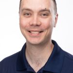 Portrait of a smiling man wearing a navy Bushnell University polo shirt against a light background.