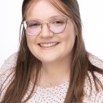Headshot of a woman with long light brown hair and glasses, wearing a light pink patterned top, smiling against a white background