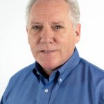 Portrait of a man with white hair wearing a blue button-down shirt against a light background.