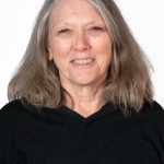 Portrait of a smiling woman with shoulder-length gray hair wearing a black top against a light background.
