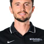 Portrait of a smiling man with short dark hair and a beard wearing a black Bushnell Soccer polo shirt against a white background