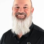 Portrait of a smiling man with a long white beard wearing a black Bushnell Athletics polo shirt against a light background.