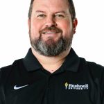 Portrait of a smiling man with a beard wearing a black Bushnell University polo shirt against a light background.
