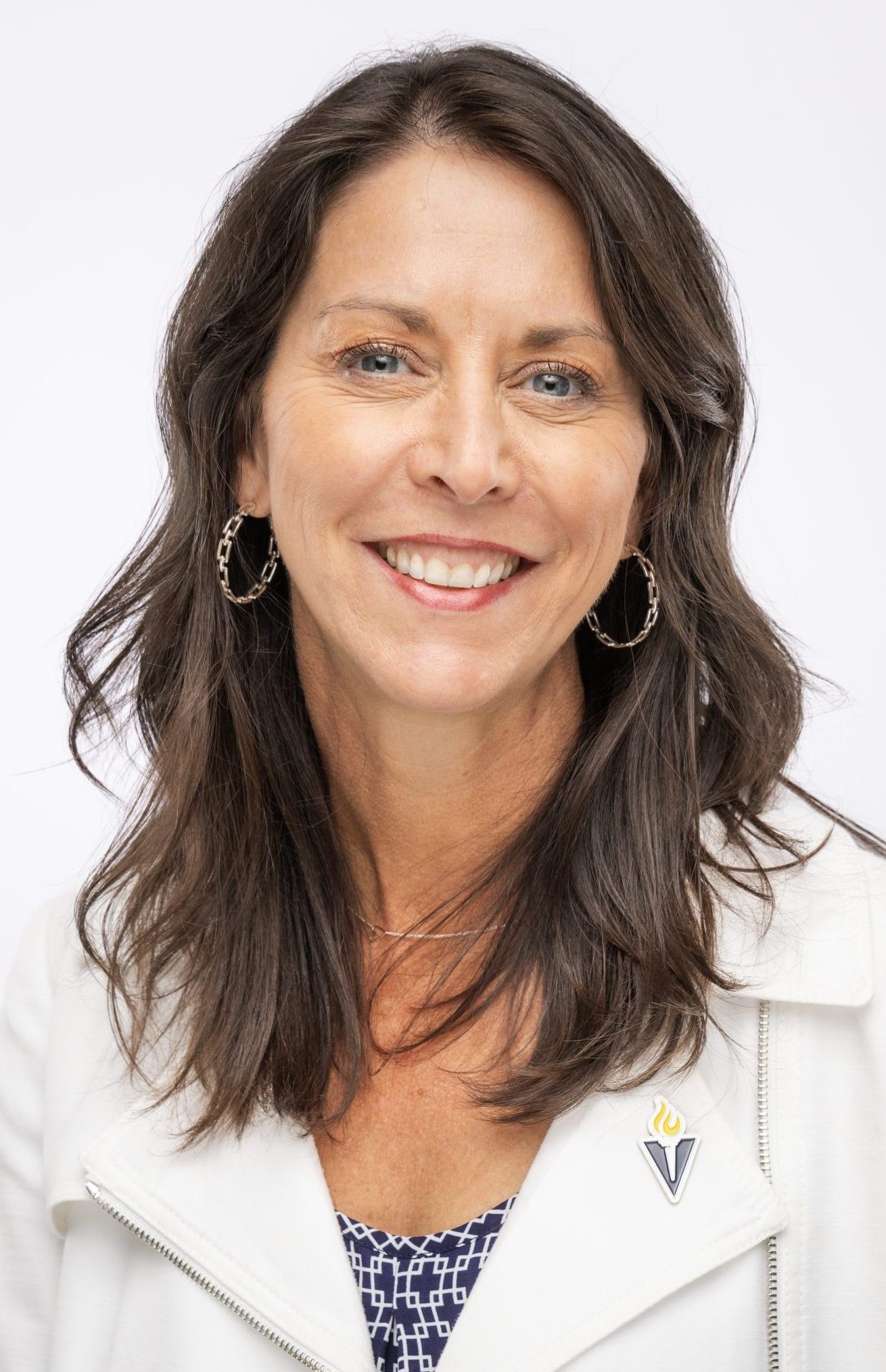Headshot of a woman with shoulder-length brown hair, wearing hoop earrings and a light-colored jacket with a Bushnell pin, smiling against a white background