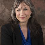 Portrait of a smiling woman with long dark hair wearing a black blazer and blue blouse against a mottled brown background.
