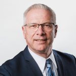 Portrait of a man with glasses wearing a navy suit and blue striped tie against a light background.