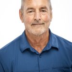 Portrait of a man with short gray hair and a trimmed beard wearing a blue button-down shirt against a light background.