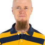 Headshot of a bald man with blue eyes and a long brown goatee wearing a yellow-and-navy striped polo shirt.