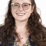 Headshot of a smiling woman with long curly brown hair and glasses wearing a denim jacket and floral top against a white background