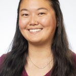 Headshot of a smiling woman with long dark hair wearing a maroon top and necklace, against a light background