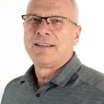 Portrait of a man wearing glasses with short gray hair and a gray Bushnell University polo shirt against a light background.