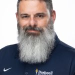Headshot of a man with short dark hair and a full gray beard wearing a navy Bushnell University polo, facing forward against a white background