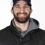 Portrait of a smiling, young man with a beard wearing a Bushnell University baseball cap, gray hoodie, and black vest against a light background.