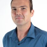 Professional headshot of a man wearing a blue button-down shirt with brown hair against a light background.