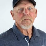 Headshot of an older man with a short white beard and blue eyes wearing a Bushnell Beacons Softball polo shirt and dark blue cap.