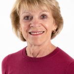 Portrait of a smiling woman with short blonde hair wearing a raspberry-colored sweater against a light background.