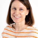 Portrait of a smiling woman with shoulder-length brown hair wearing a cream-colored shirt with orange stripes against a light background.
