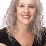 Headshot of a smiling woman with curly gray hair wearing a black top and a necklace, facing forward against a white background
