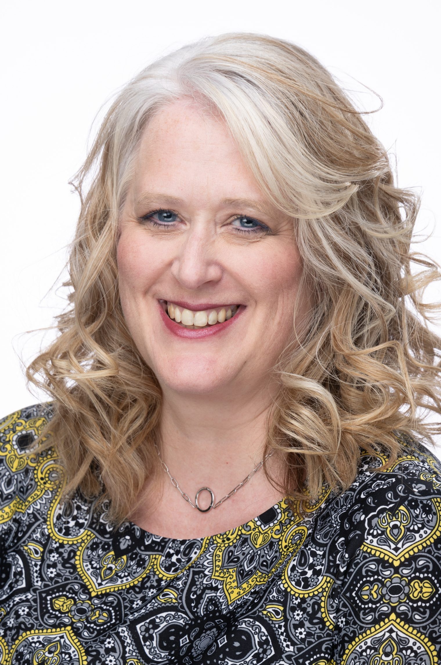 Headshot of a woman with shoulder-length blonde curly hair, wearing a patterned black and yellow top and a necklace, smiling against a white background