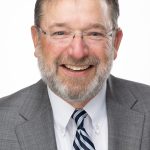 Headshot of a smiling man with glasses and a gray beard wearing a suit and striped tie against a white background