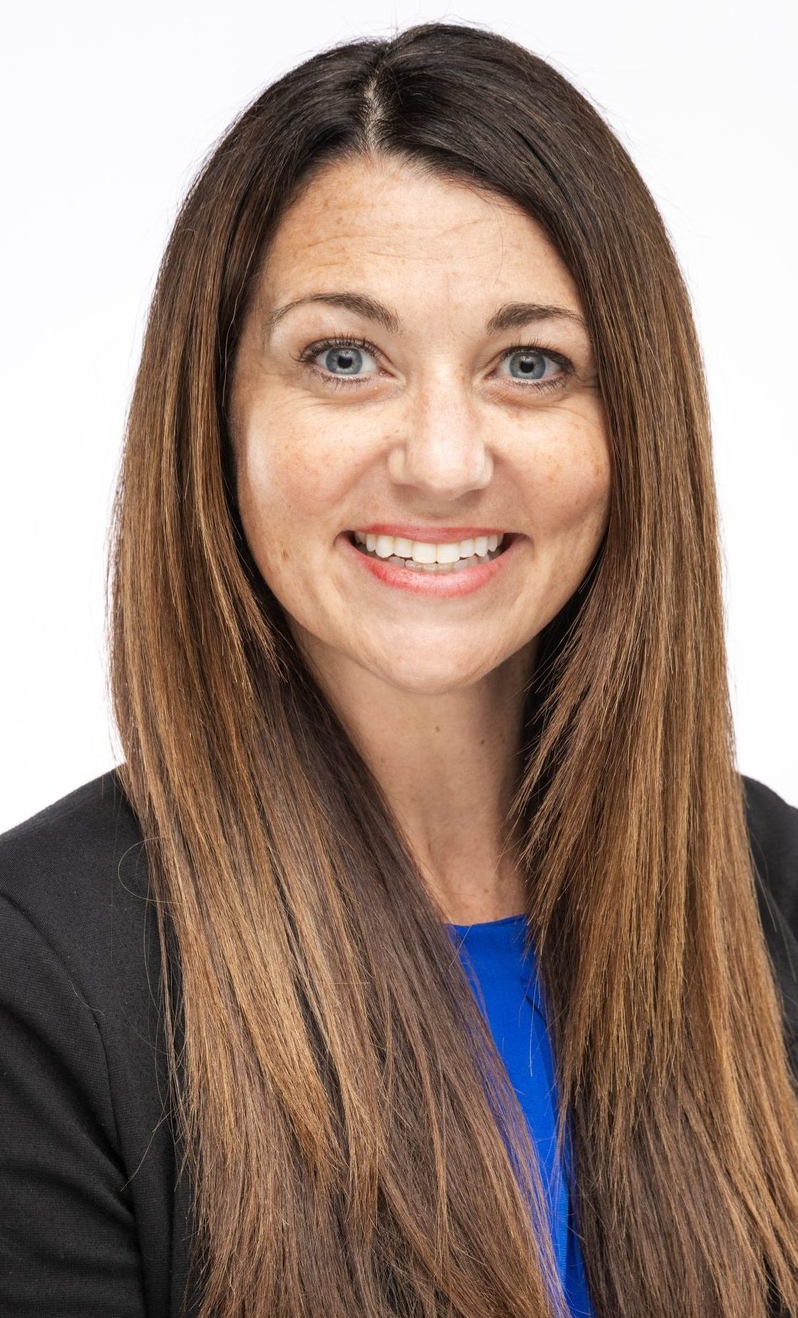 Headshot of a woman with long straight brown hair, wearing a black blazer and blue top, smiling against a white background