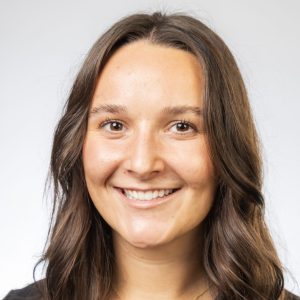 Headshot of a smiling woman with long dark hair wearing a black top and necklace, against a light background.