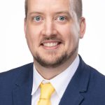Headshot of a man with short light brown hair and a trimmed beard, wearing a navy suit and yellow tie, smiling against a white background