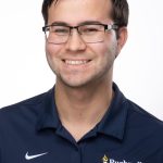 Headshot of a smiling man with short dark hair and glasses wearing a navy Bushnell University polo shirt against a white background