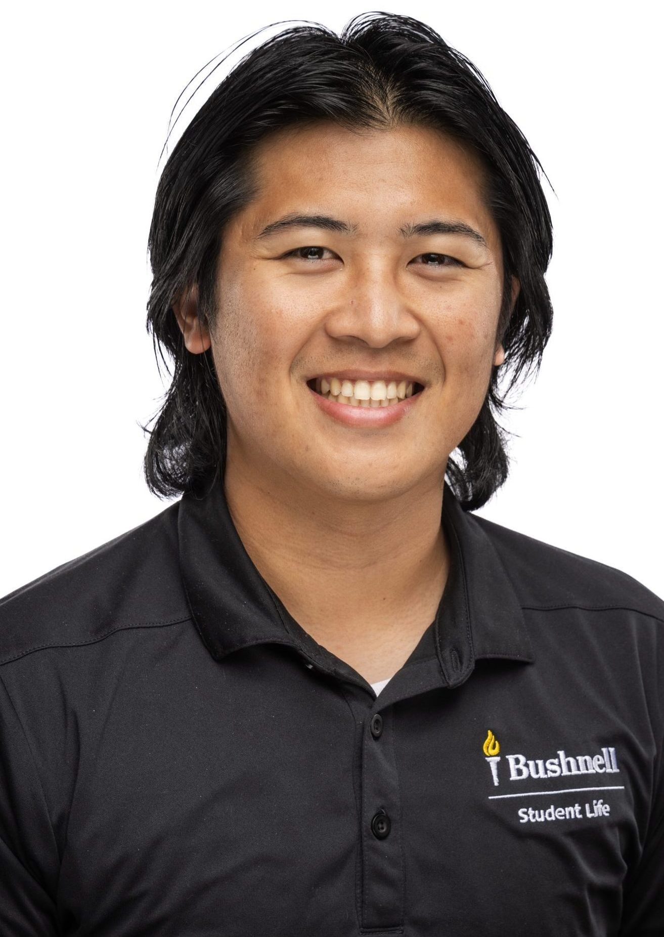 Headshot of a man with medium-length dark hair, wearing a black Bushnell Student Life polo shirt, smiling against a white background