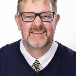 Headshot of a man with short light brown hair, glasses, and a trimmed beard, wearing a navy sweater and striped tie, smiling against a white background