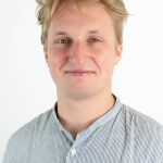 Portrait of a young man with blond hair wearing a light striped button-up shirt against a light background.