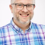 Headshot of a man with short gray hair, glasses, and a trimmed beard, wearing a blue and purple plaid button-up shirt, smiling against a white background