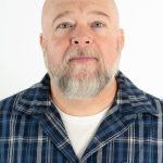 Portrait of a man with a gray beard wearing a blue plaid shirt against a light background.
