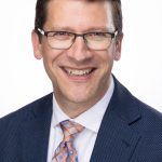 Headshot of a man with short brown hair and glasses wearing a blue suit and patterned tie, smiling against a white background