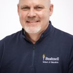 Professional headshot of a smiling bald man with a gray goatee wearing a navy Bushnell School of Education pullover.