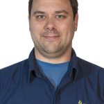 Headshot of a man with short dark hair and light facial stubble wearing a blue shirt with a Bushnell University logo against a white background