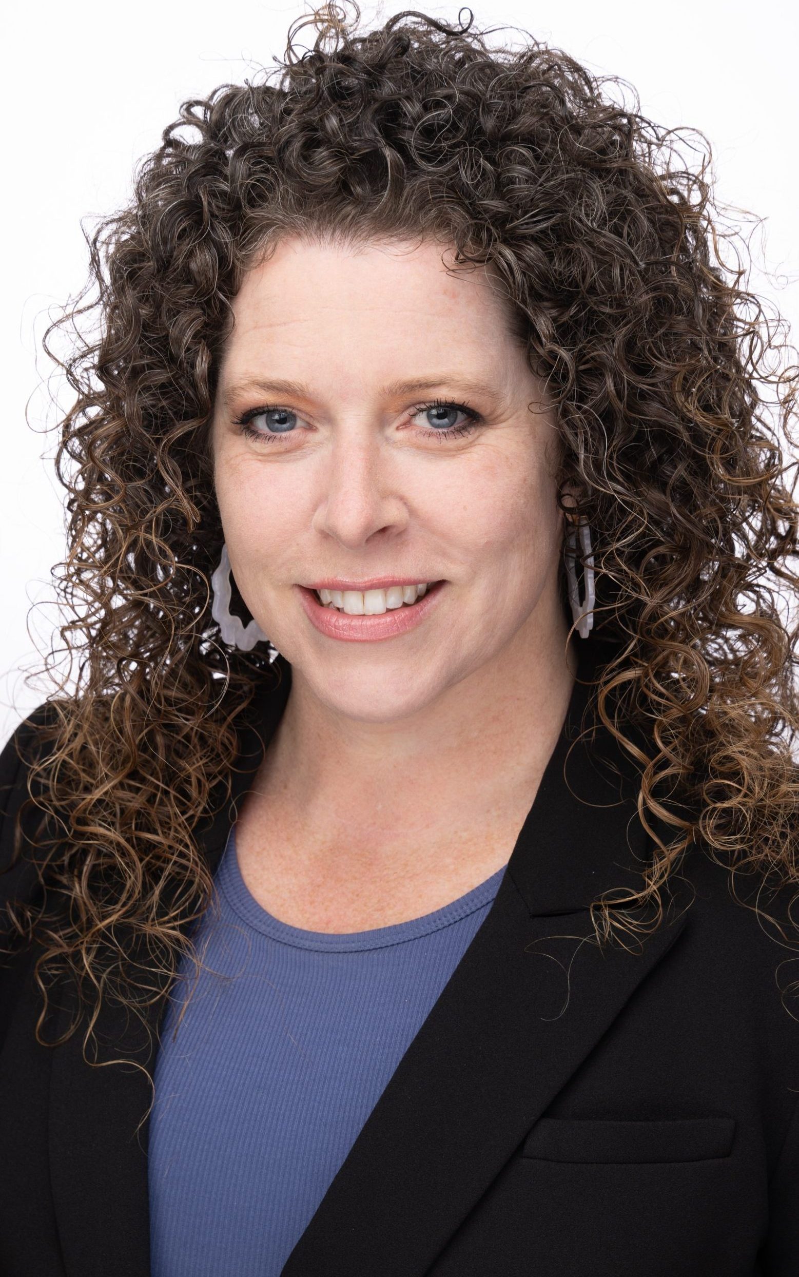 Headshot of a woman with long curly brown hair, wearing a black blazer and blue top, smiling against a white background