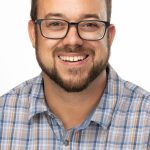 Headshot of a man with short dark hair, glasses, and a trimmed beard, wearing a plaid button-up shirt and smiling against a white background