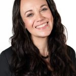 Portrait of a smiling woman with long dark wavy hair wearing a black top against a light background.