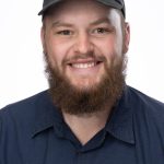 Headshot of a smiling man with a beard wearing a black Bushnell University cap and navy shirt, facing forward against a white background