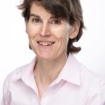 Portrait of a smiling woman with short brown hair wearing a light pink button-up shirt against a light background.