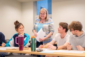 Professor standing and speaking with students seated around a classroom table with papers and notebooks