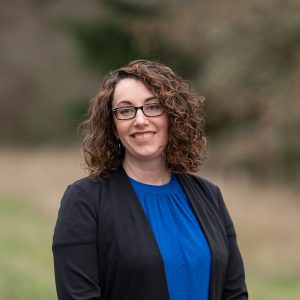Portrait of a smiling woman with curly brown hair and glasses wearing a black blazer over a blue blouse, standing outdoors against a softly blurred background.
