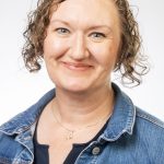 Headshot of a woman with short curly light brown hair, wearing a denim jacket and necklace, smiling against a white background