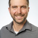Headshot of a man with short light brown hair and a trimmed beard, wearing a gray button-up shirt, smiling against a white background