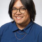 Headshot of a smiling person with medium-length dark hair and glasses, wearing a blue button-down shirt and gold necklace against a white background