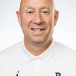 Headshot of a smiling bald man with light stubble, wearing a white Bushnell Women’s Basketball polo against a white background
